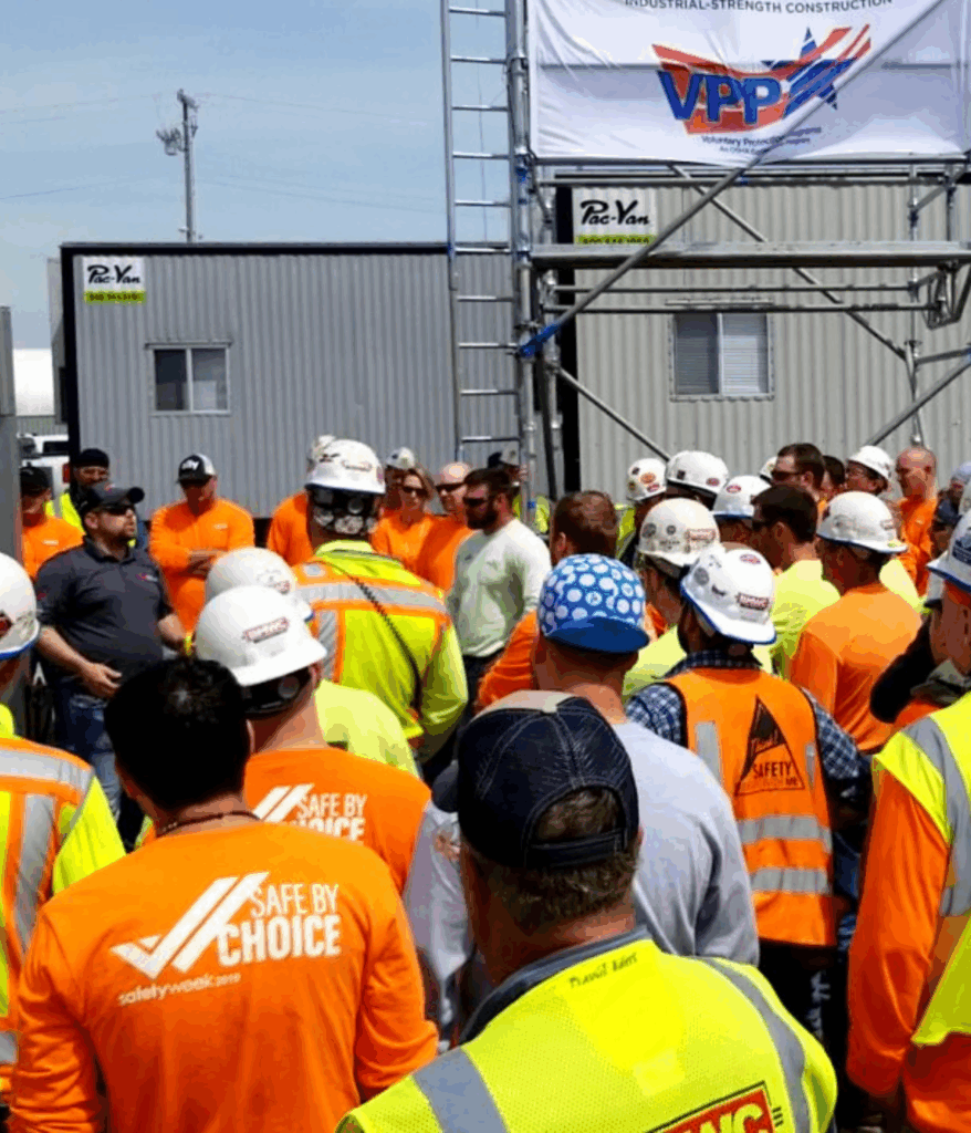 Group of construction workers watching speaker VPP signage BMWC logo HiViz Safety Yellow long sleeve shirts HiViz safety orange long sleeve shirts  HiViz safety vests with reflective strips hard hats 